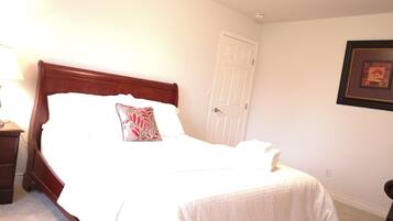 A neatly made bed with a white comforter and a decorative pillow, a wooden nightstand with a lamp, and a framed picture on the wall.