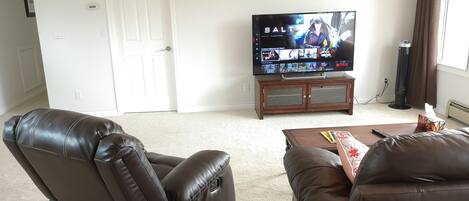 A living room with a flat-screen TV on a wooden stand, leather reclining chairs, a coffee table with remote controls, and a clock on the wall.