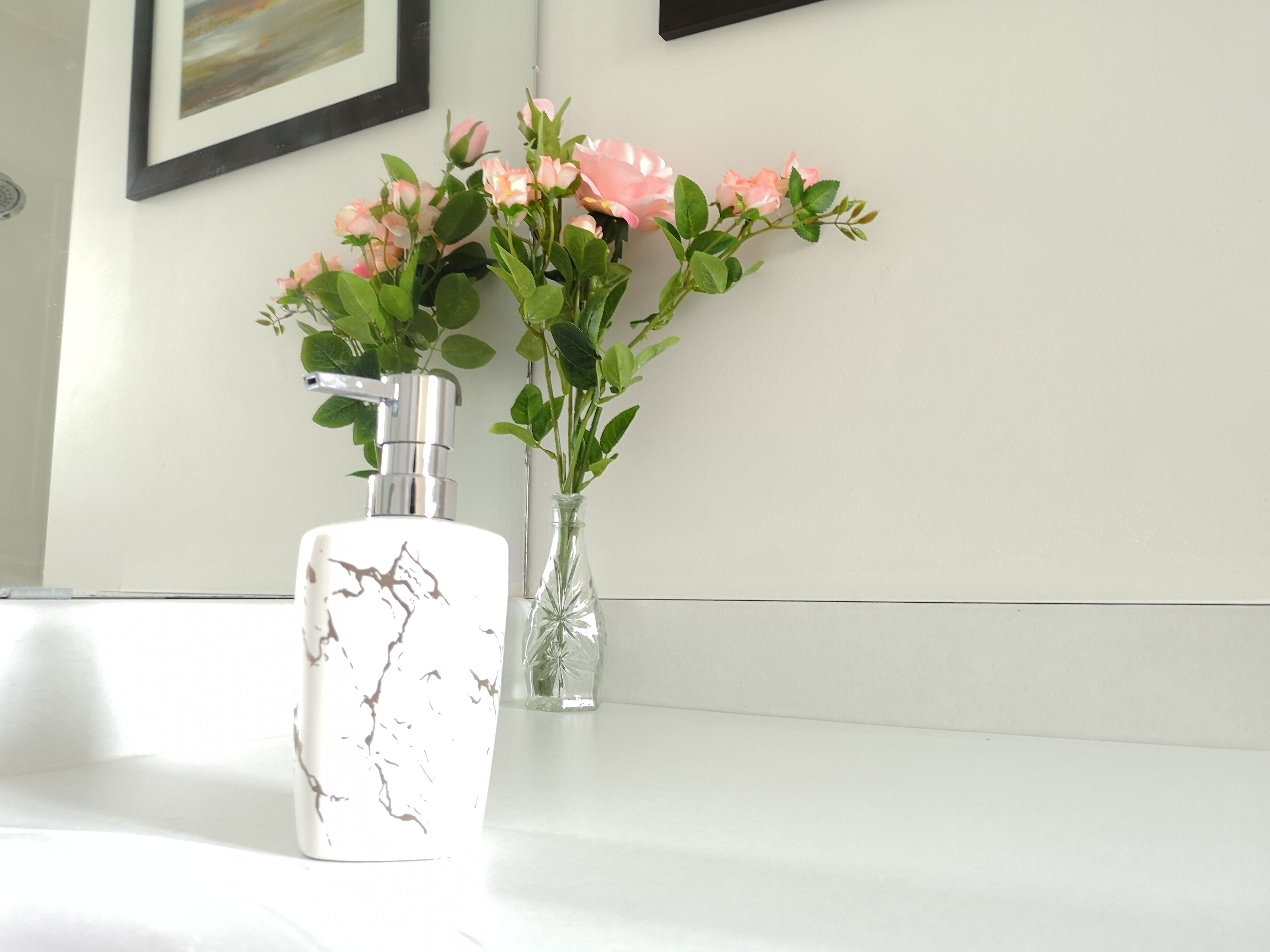 Un baño con lavamanos blanco, una botella con diseño de mármol y un florero con flores rosas.
