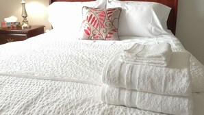 Una cama bien hecha con sábanas blancas y una almohada decorativa, cabecera de madera y una mesita de noche con una lámpara y una caja de pañuelos.