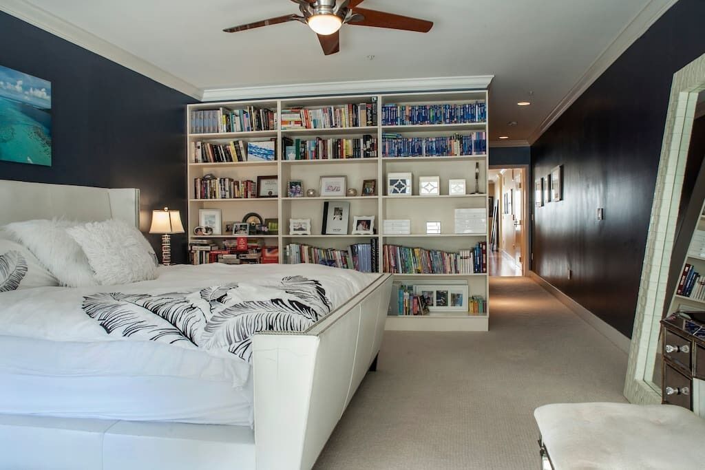 A bedroom with a large bed, a bookshelf, a ceiling fan, and a dark wall.