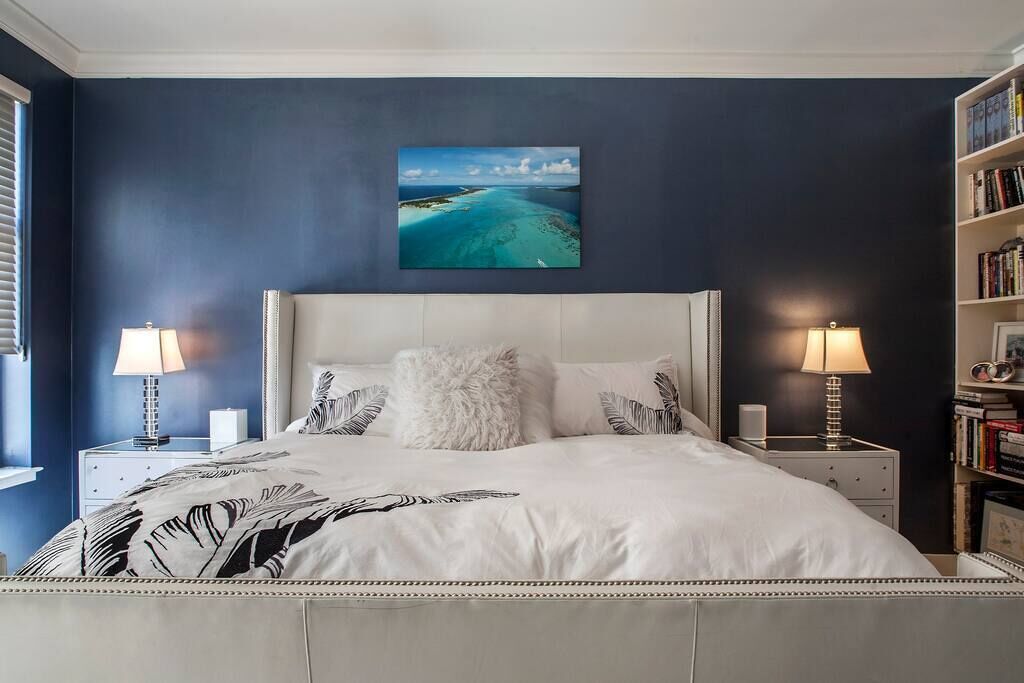 A bedroom with a large bed, two bedside tables with lamps, a bookshelf, and a painting of a coastal landscape on the wall.