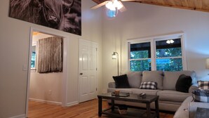 A living room with a wooden ceiling, a ceiling fan, a large window with a view of trees, a grey sofa with cushions, and a coffee table.