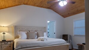 A bedroom with a large bed, a nightstand, a lamp, a wooden ceiling, and a window with blinds.