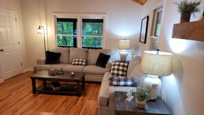 A living room with a sofa, coffee table, and lamps. There are windows with a view of trees.