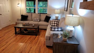 A living room with a sofa, coffee table, and lamps. There are windows with a view of trees.