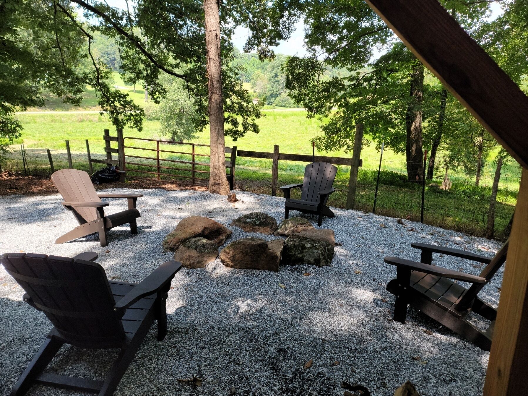 An outdoor seating area with Adirondack chairs and a wooden table, surrounded by a gravel ground and a wooden fence.