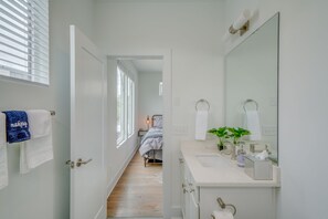 Ein schmaler Flur führt zu einem Schlafzimmer mit Bett und Nachttisch. Rechts ist ein Badezimmer mit Waschbecken, Spiegel und Handtuchhalter zu sehen.