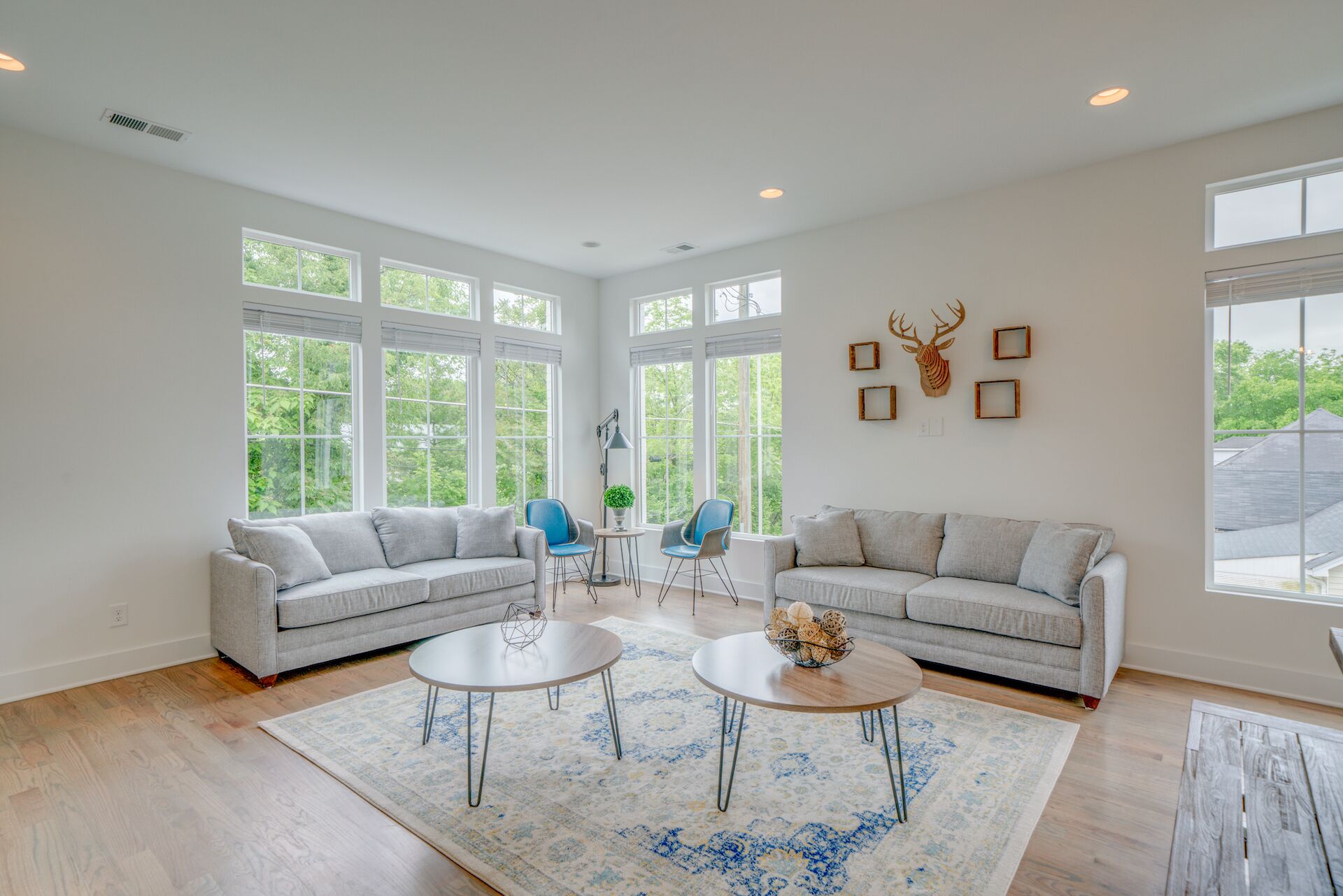 A spacious living room with two grey sofas, a coffee table, and a blue chair. Large windows allow natural light and views of greenery.