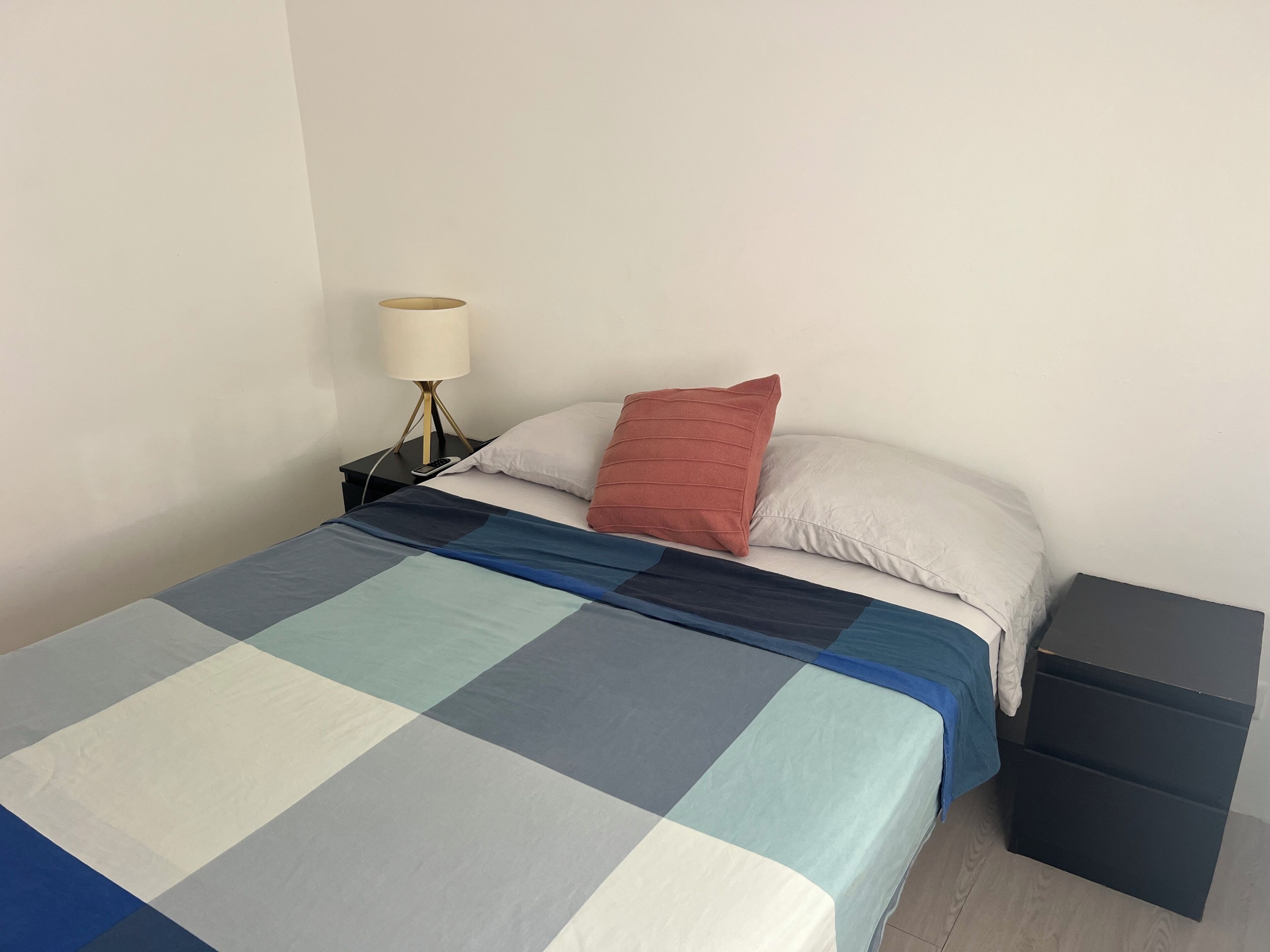 Ein Bett mit kariertem Überwurf, ein Nachttisch mit Lampe und eine kleine, schwarze Aufbewahrungsbox.