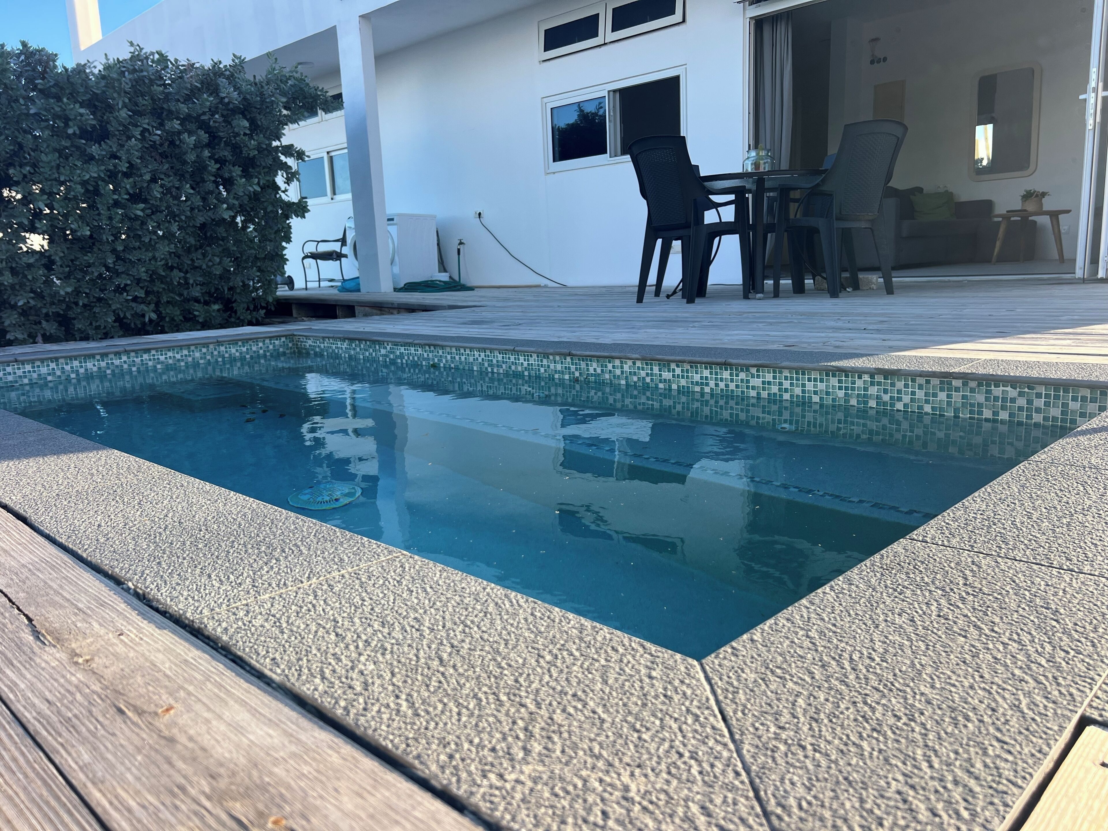 A swimming pool with a tiled edge, surrounded by a patio area with a table and chairs, and a hedge in the background.