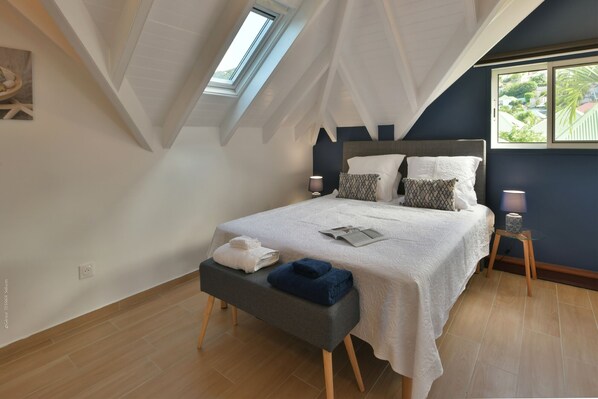 A bedroom with a sloped ceiling, a bed with white bedding, a bedside table with a lamp, and a window with a view of greenery.