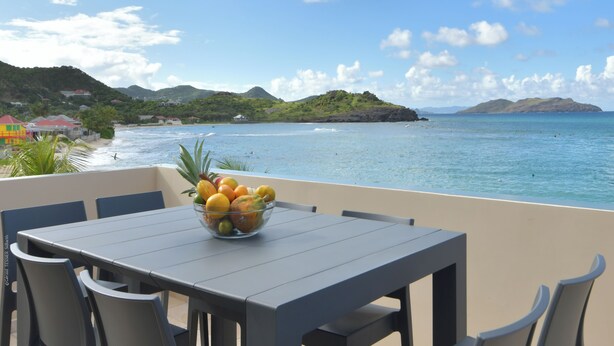 Un balcon avec une table et des chaises, un panier de fruits et une vue sur la mer et les collines.