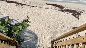 Un escalier en bois mène à une plage de sable fin, bordée d’une eau d’un bleu limpide et dominée par des collines lointaines.