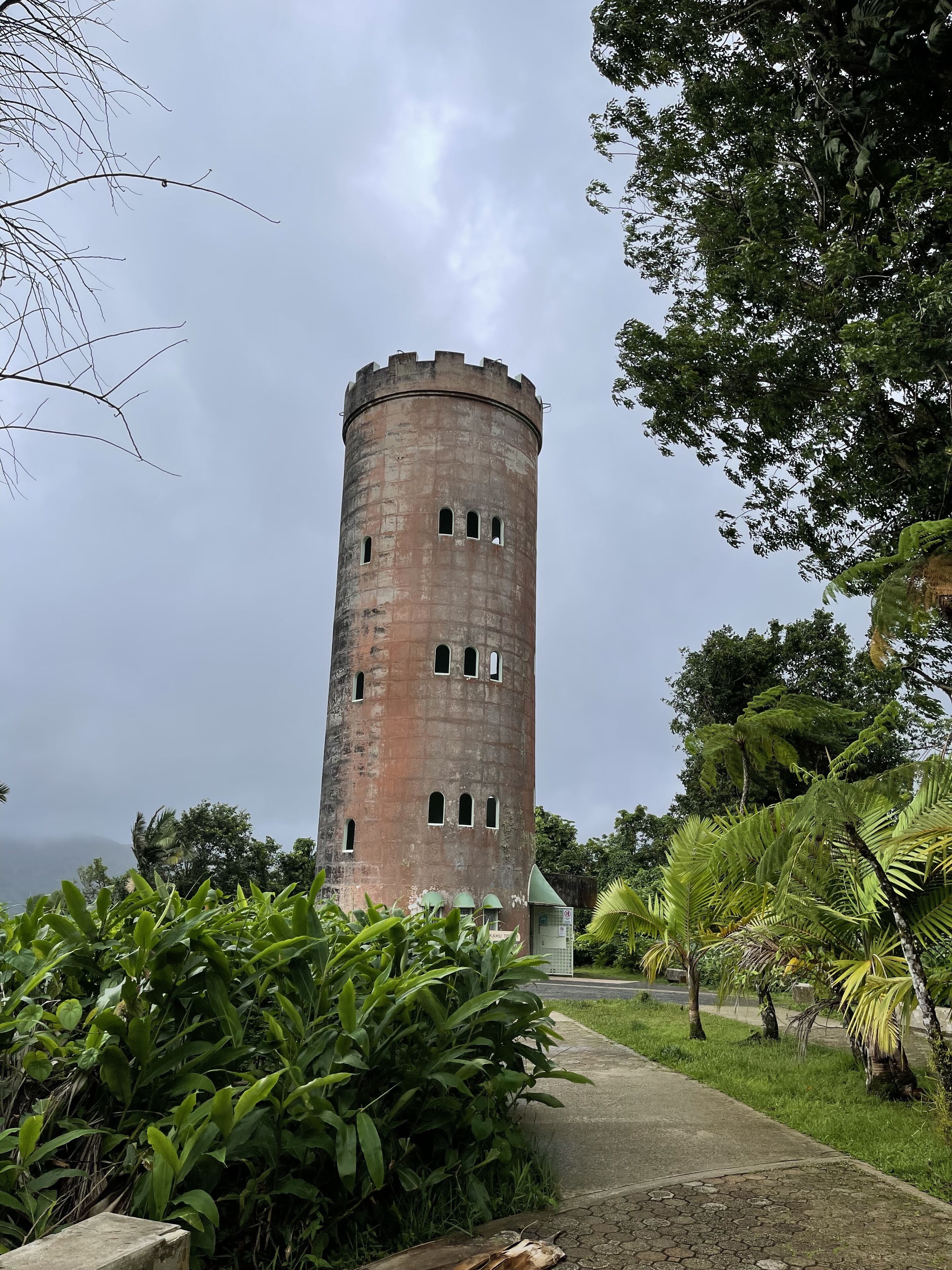 A historic tower with a brick exterior and multiple windows, surrounded by lush greenery and a pathway leading to it.