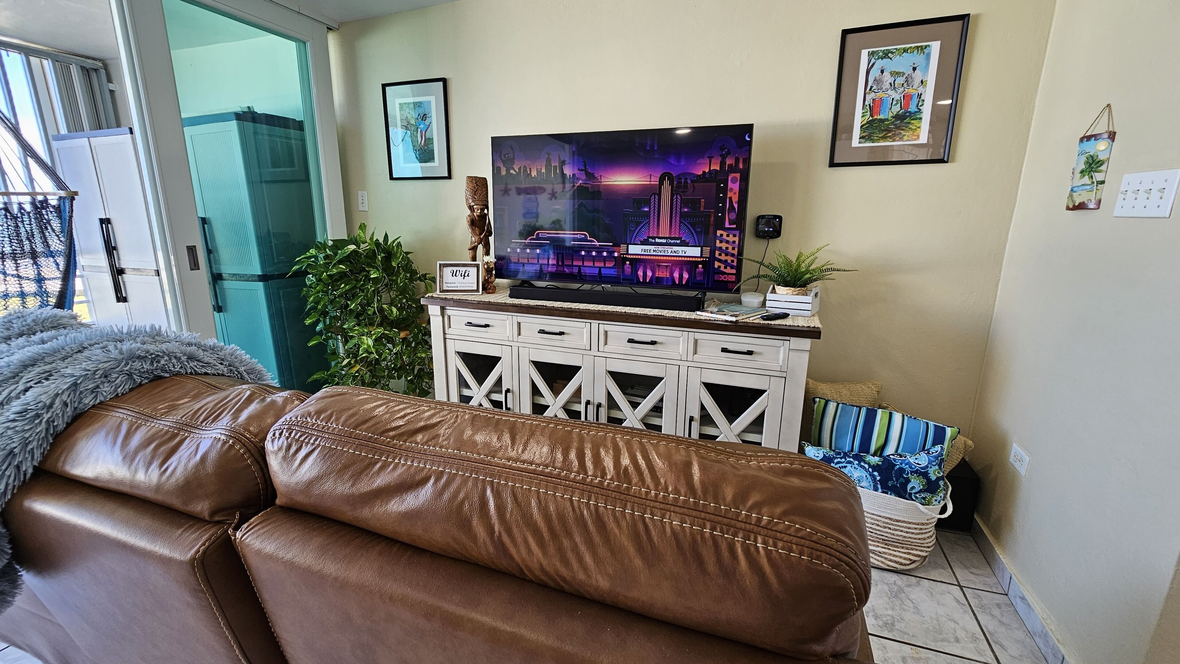 Un salon avec un canapé en cuir, un téléviseur posé sur un meuble blanc et une porte donnant sur l’extérieur.
