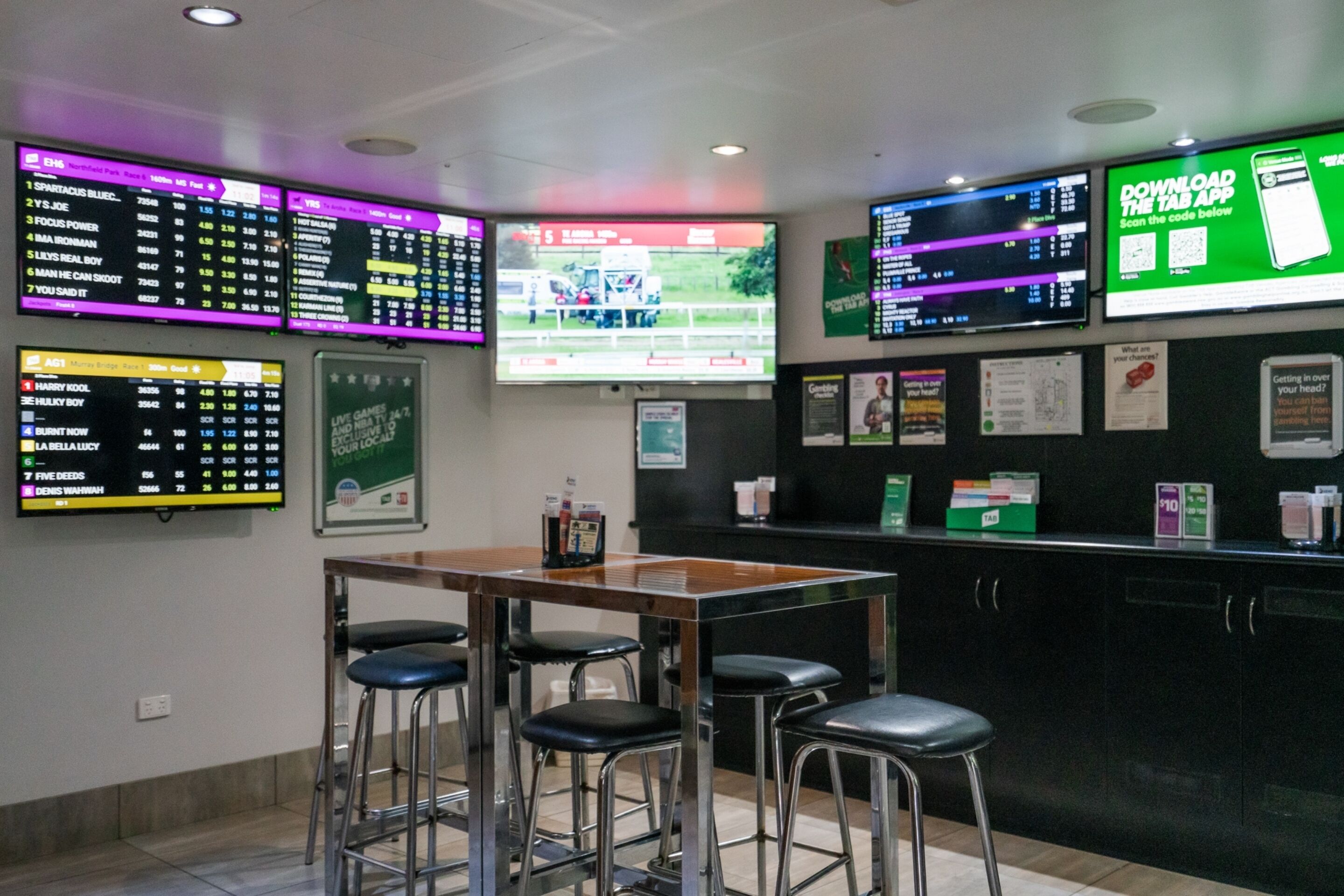 A bar area with multiple screens displaying sports betting information, a wooden table, and bar stools.