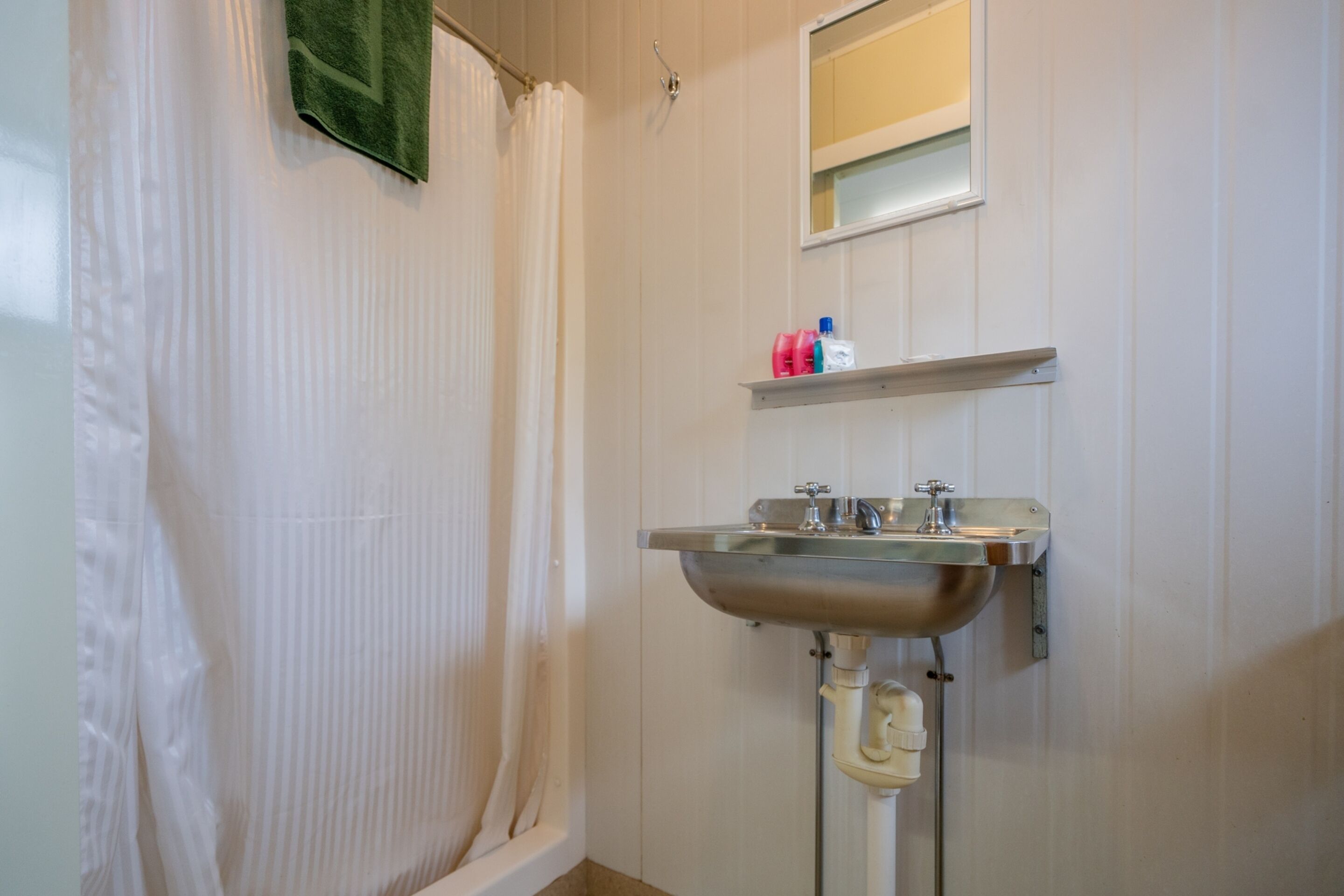 A bathroom with a white shower curtain, a sink with a mirror above it, and a shelf with toiletries.