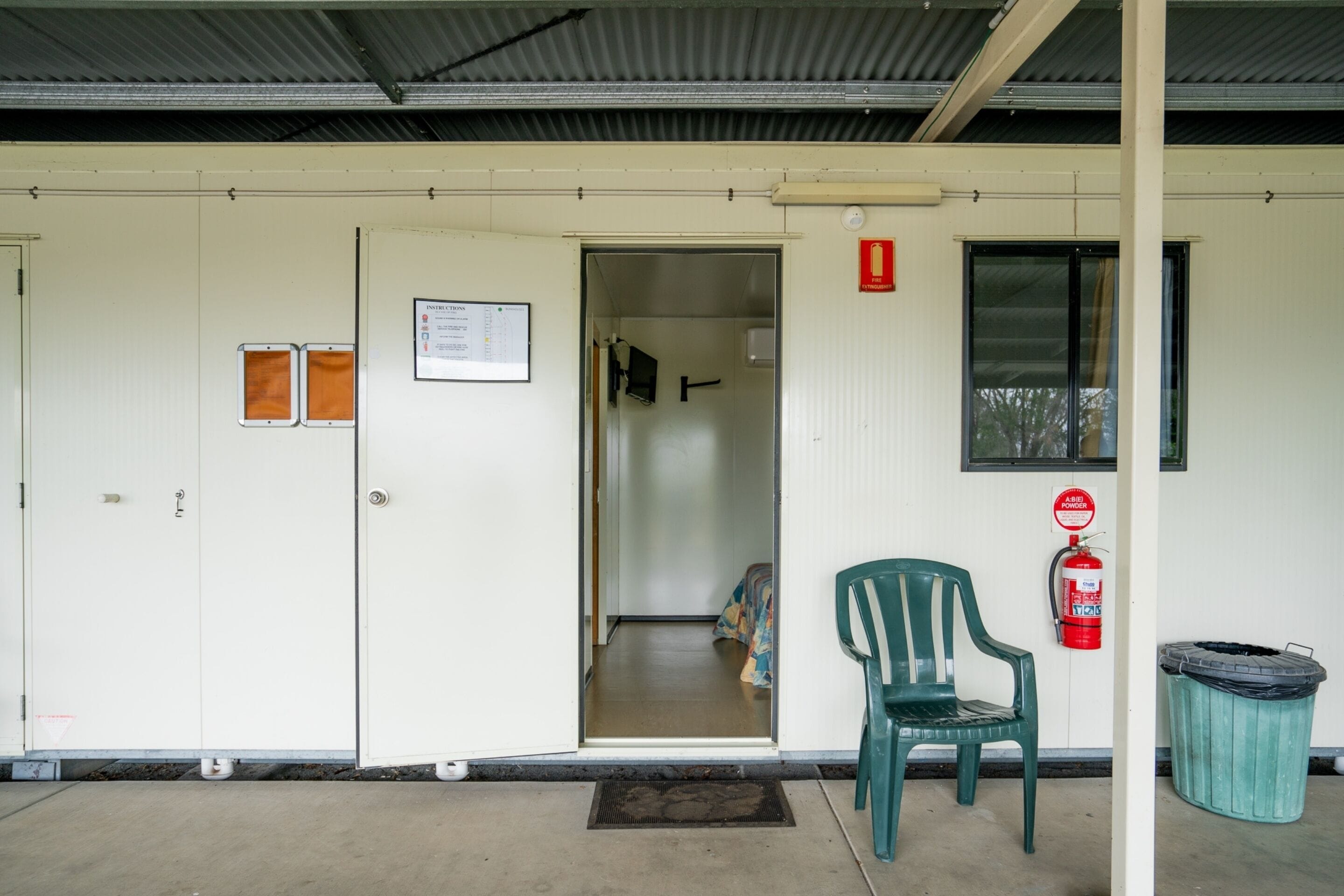 A building entrance with a door, a fire extinguisher, a trash bin, and a chair.