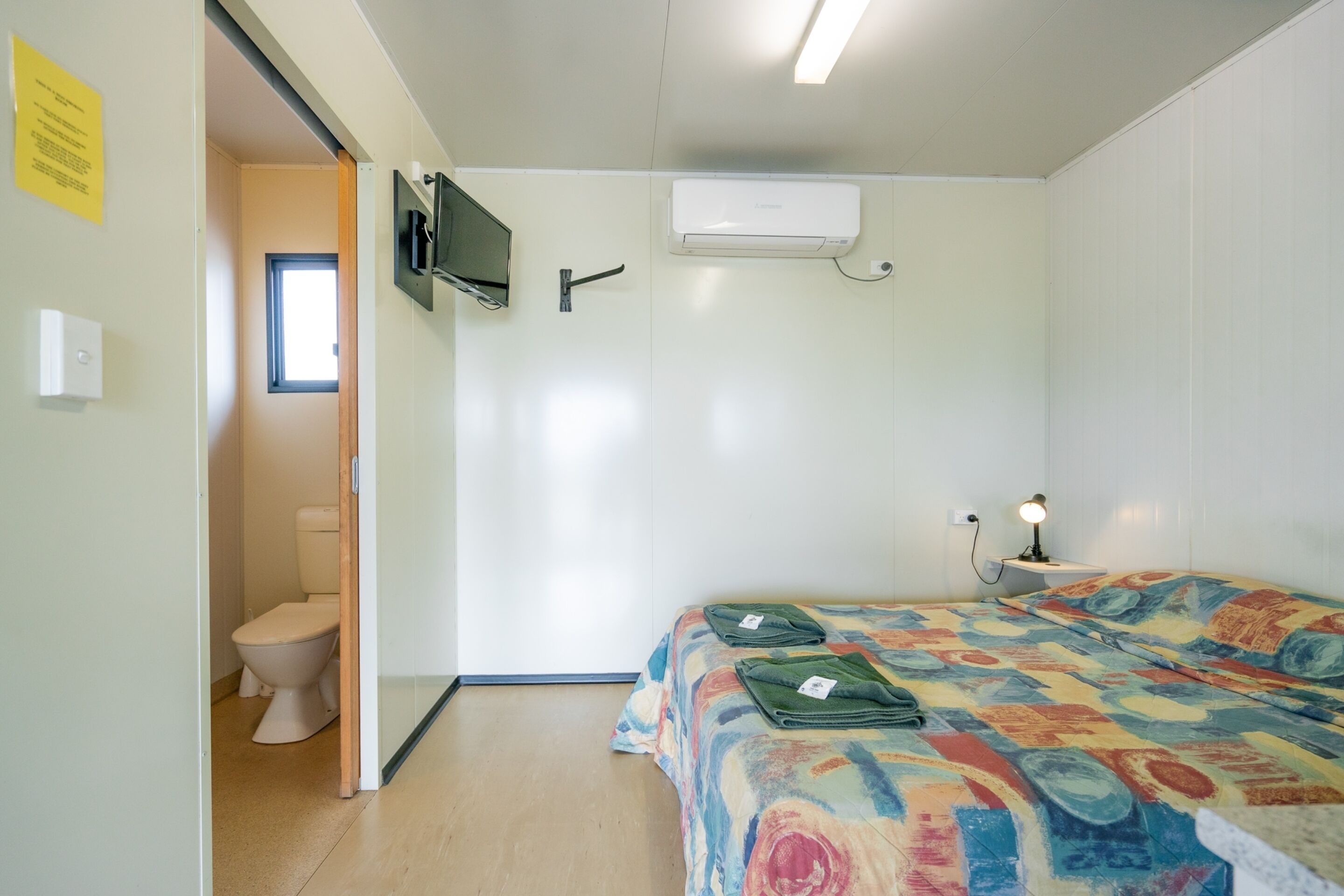 A bedroom with a bed, an air conditioner, a TV mounted on the wall, and a window.