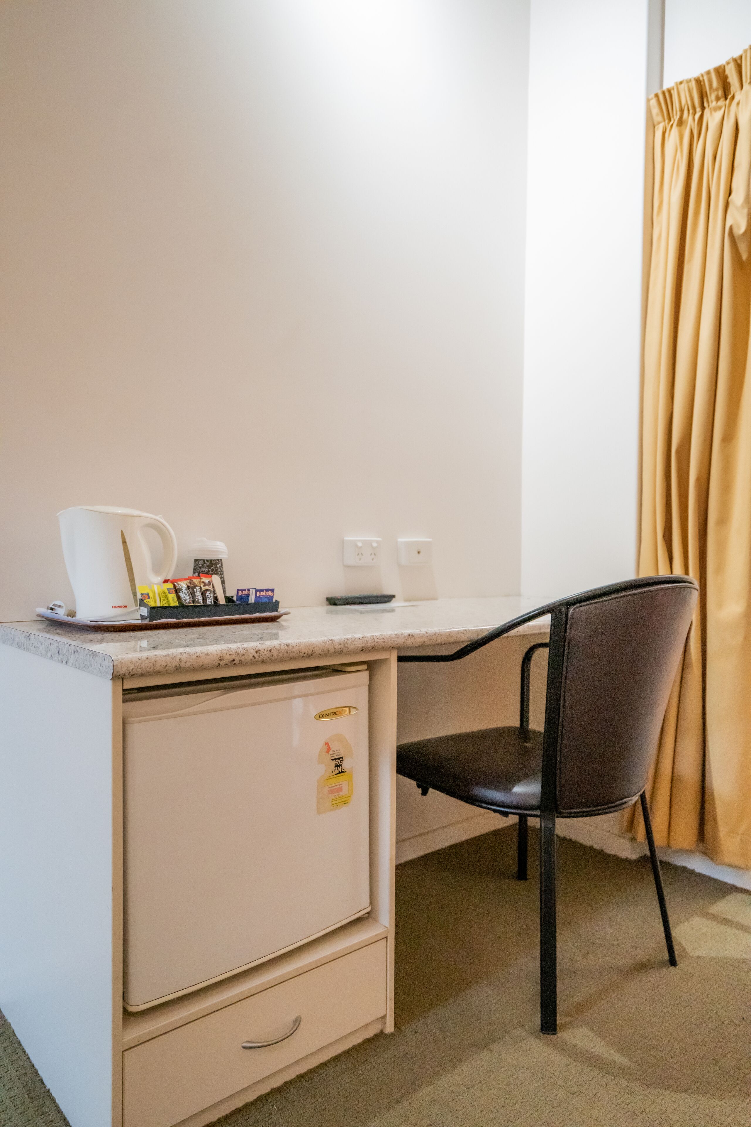 A small kitchenette with a coffee maker, a kettle, and a microwave. There is a chair and a cabinet below the counter.