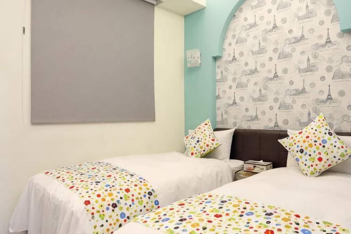 A room with two beds, a large window, a patterned headboard, and a grey wall.