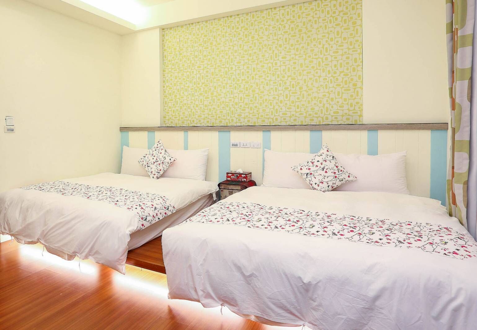 A hotel room with two single beds, a wooden floor, a patterned wall, and a curtain on the right.