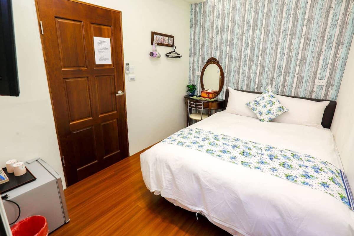 A bedroom with a wooden door, a desk with a chair, a bed with a floral bedspread, a bedside table with a mirror, and a striped curtain.