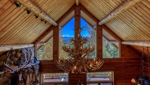 A log cabin interior with a large chandelier made of antlers, stone walls, and a view of mountains through the windows.