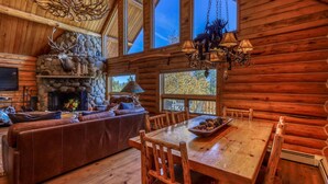 A log cabin interior with a stone fireplace, leather sofa, dining table, and large windows.