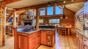 A log cabin interior with a stone fireplace, wooden dining area, and large windows.