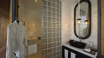 Un baño con cabina de ducha de vidrio, pared de azulejos blancos, lavamanos negro y espejo redondo, con una bata colgada en un perchero.