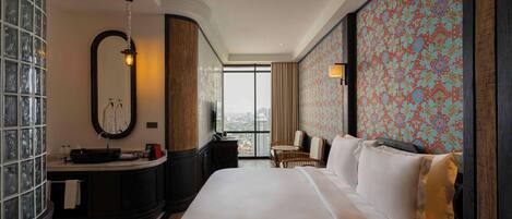 A hotel room with a large bed, a vanity with a mirror, a small table, and a view of the city through the window.