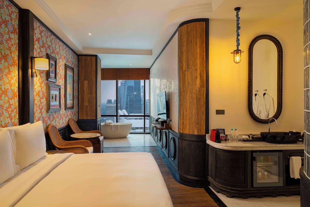 Een hotelkamer met een groot bed, een zithoek met stoelen, een badkuip, een wastafel met spiegel en uitzicht op de stad.