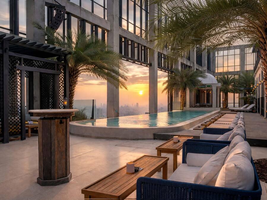 A rooftop pool area with lounge seating and palm trees, overlooking a cityscape at sunset.