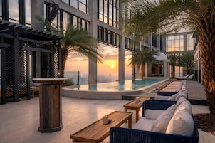 Un área de piscina en la azotea con mobiliario de descanso y palmeras, con vistas a un paisaje urbano al atardecer.