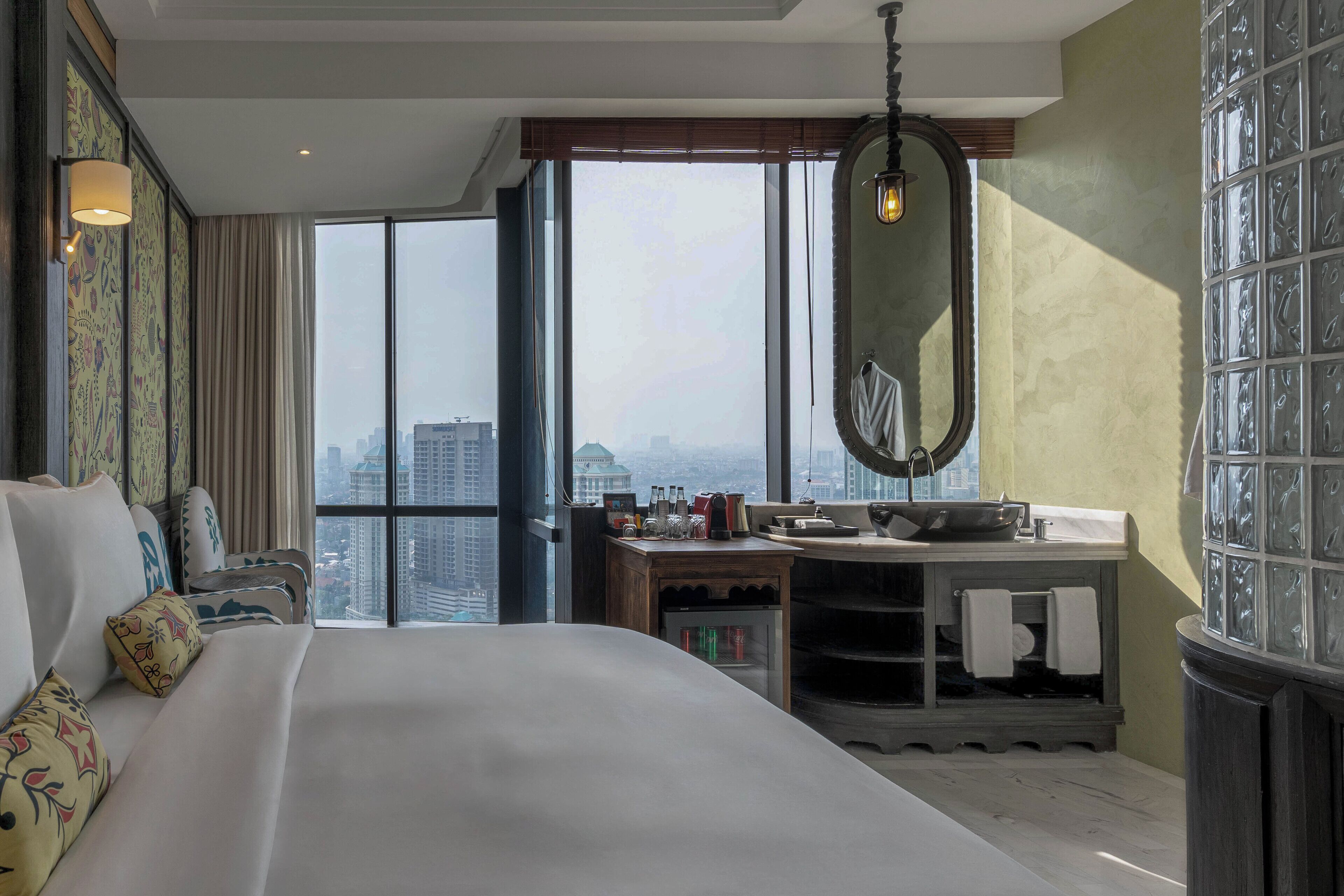 A hotel room with a large bed, a vanity with a mirror, and a view of the city through large windows.