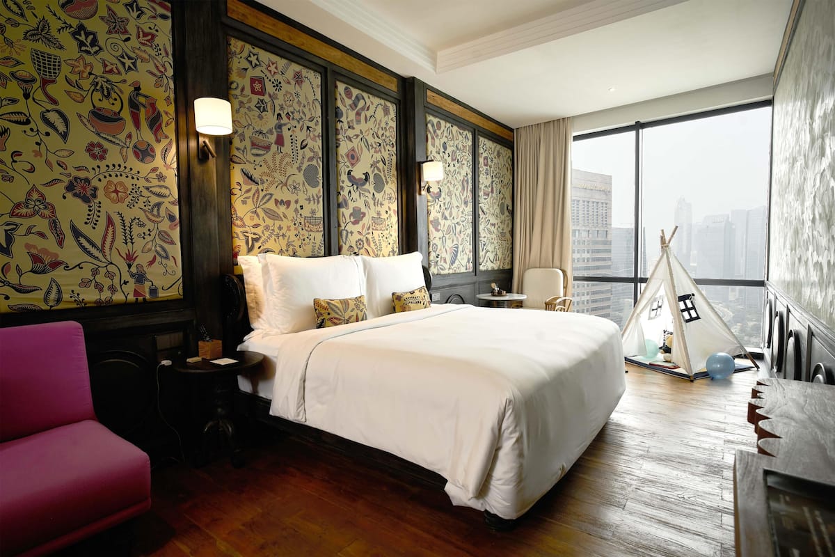 Een hotelkamer met een groot bed, een roze stoel, een klein tafeltje, een lamp, een tipi en uitzicht op de stad.