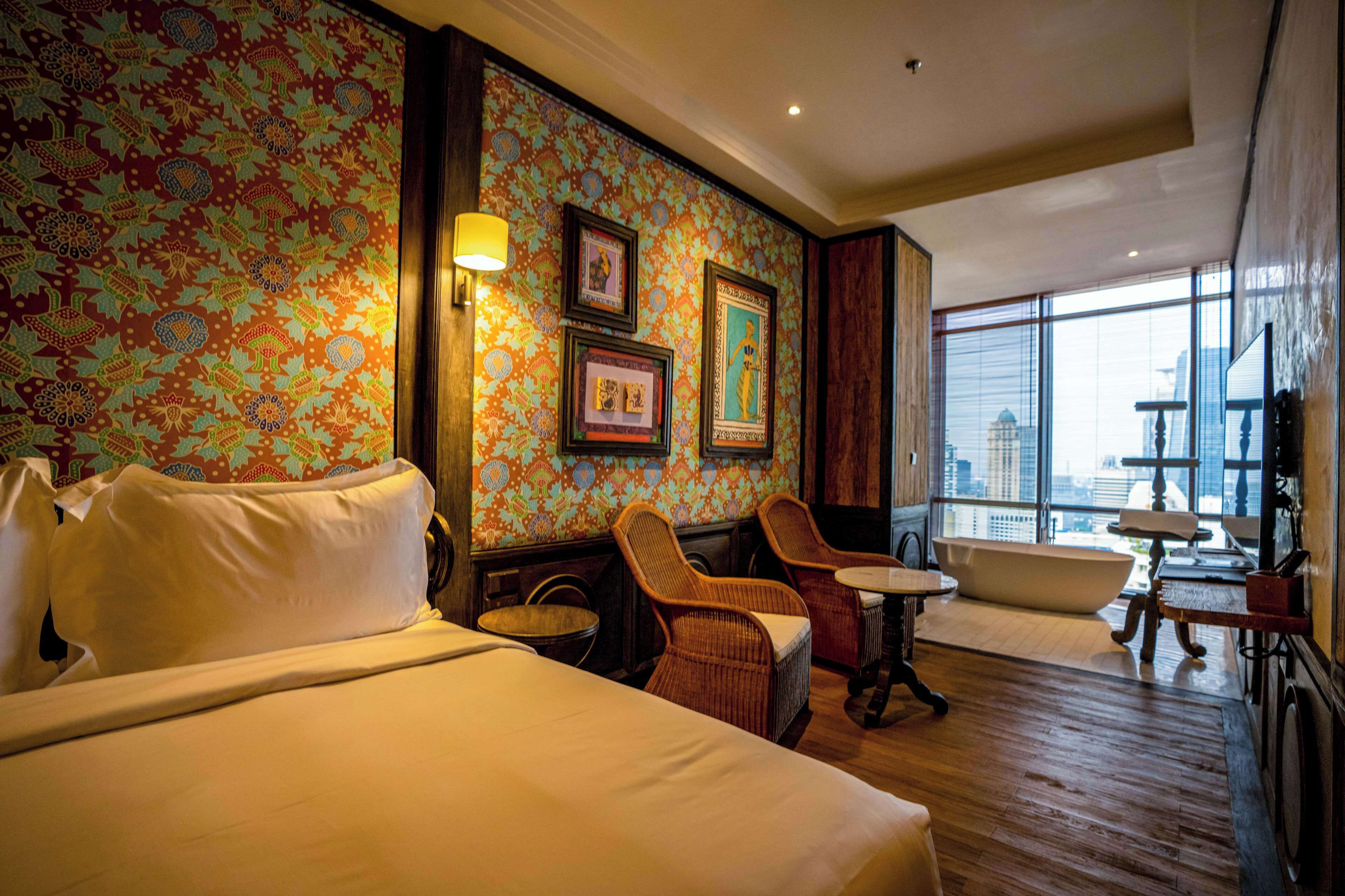 A hotel room with a large bed, two wicker chairs, a small table, and a large window offering a city view.