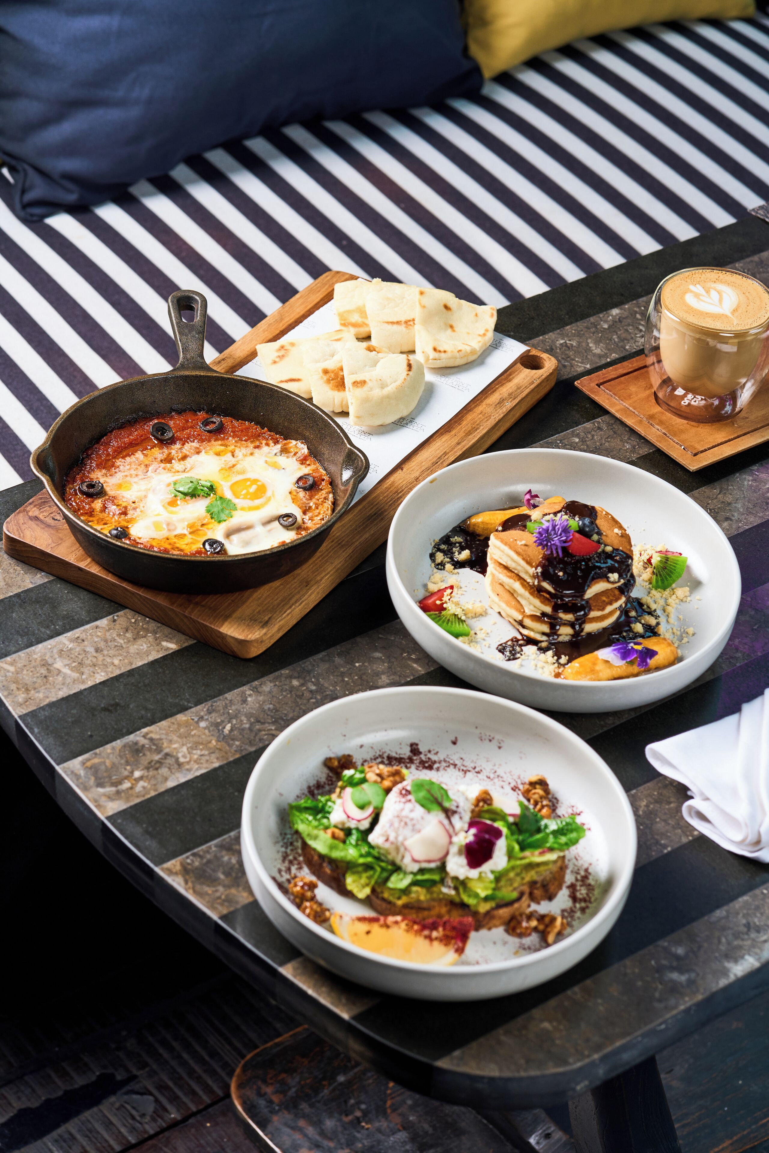 Una mesa de comedor con tres platos: una sartén con huevos y aceitunas, una pila de tortitas con salsa de chocolate y fruta, y un bol con ensalada.