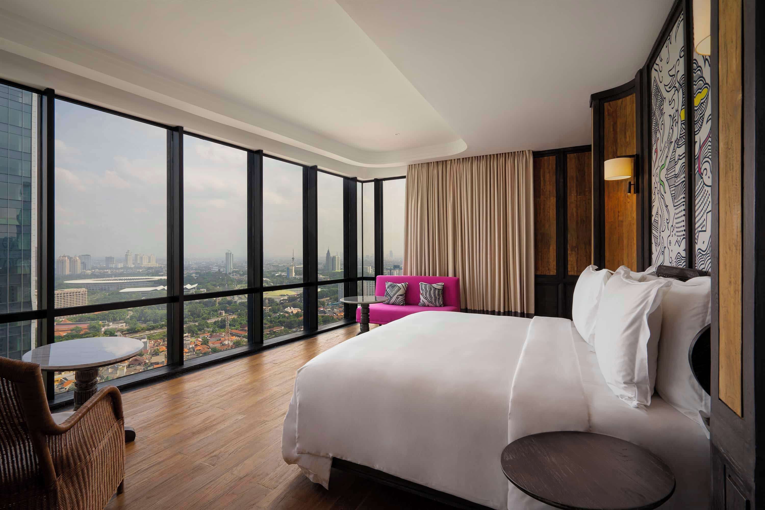 A hotel room with a large bed, a small table, a chair, and a view of the city through large windows.