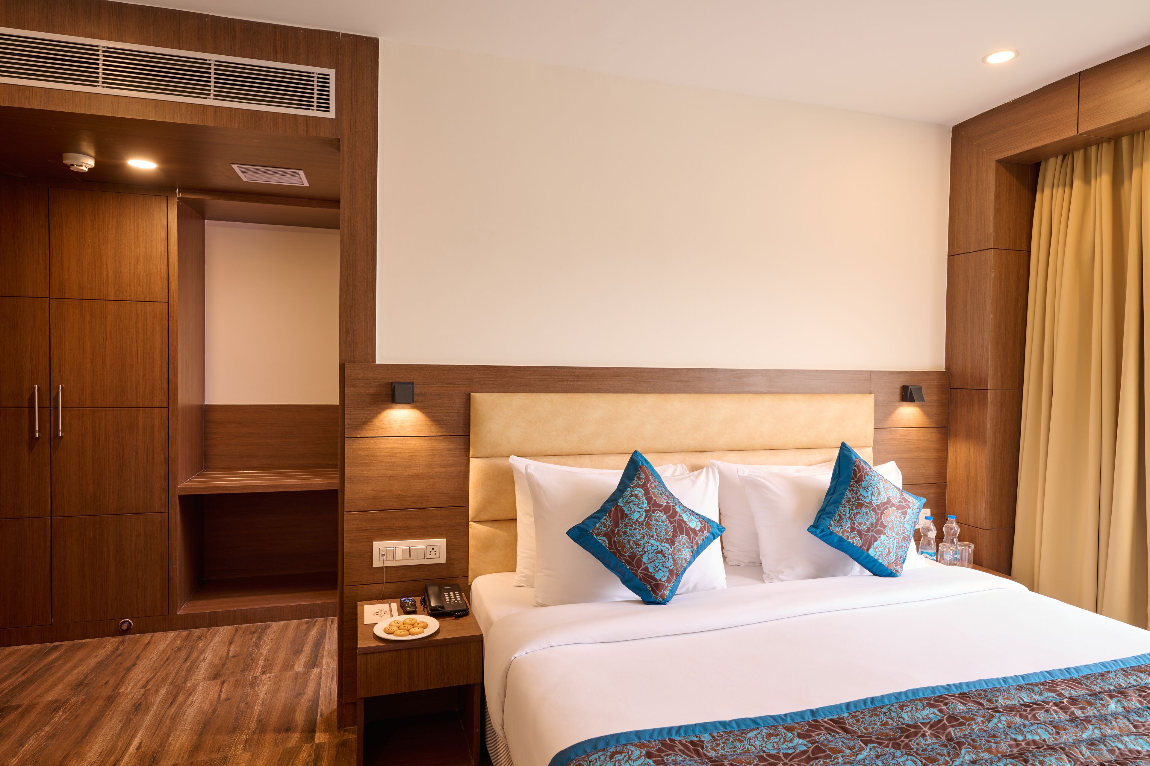 A hotel room with a large bed, two pillows, a nightstand with a phone and a plate of cookies, a wooden wardrobe, and a headboard with built-in lighting.