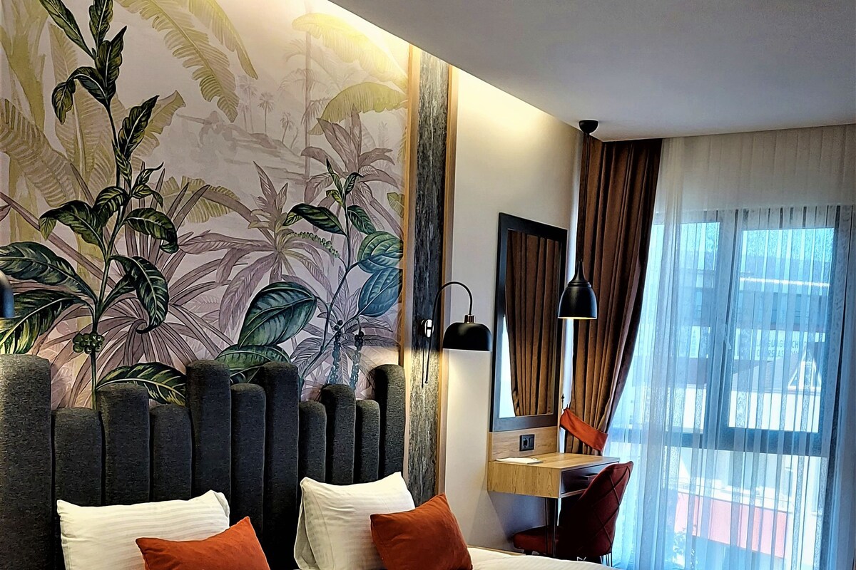 A modern hotel room with a large bed, a desk, and a view of the city through sheer curtains.