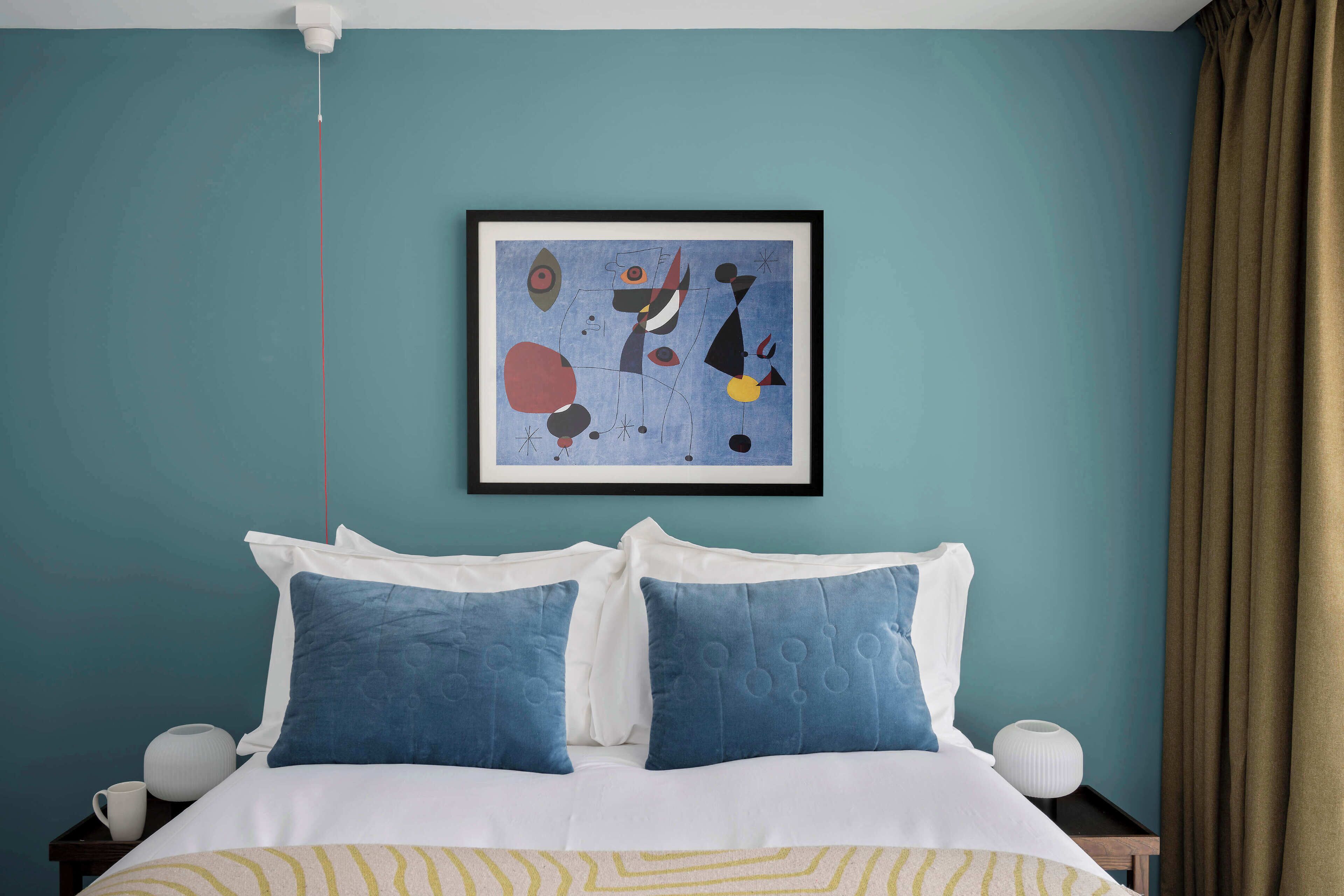 Una cama bien hecha con ropa de cama azul y blanca, un cuadro abstracto enmarcado en la pared y una cortina marrón a la derecha.
