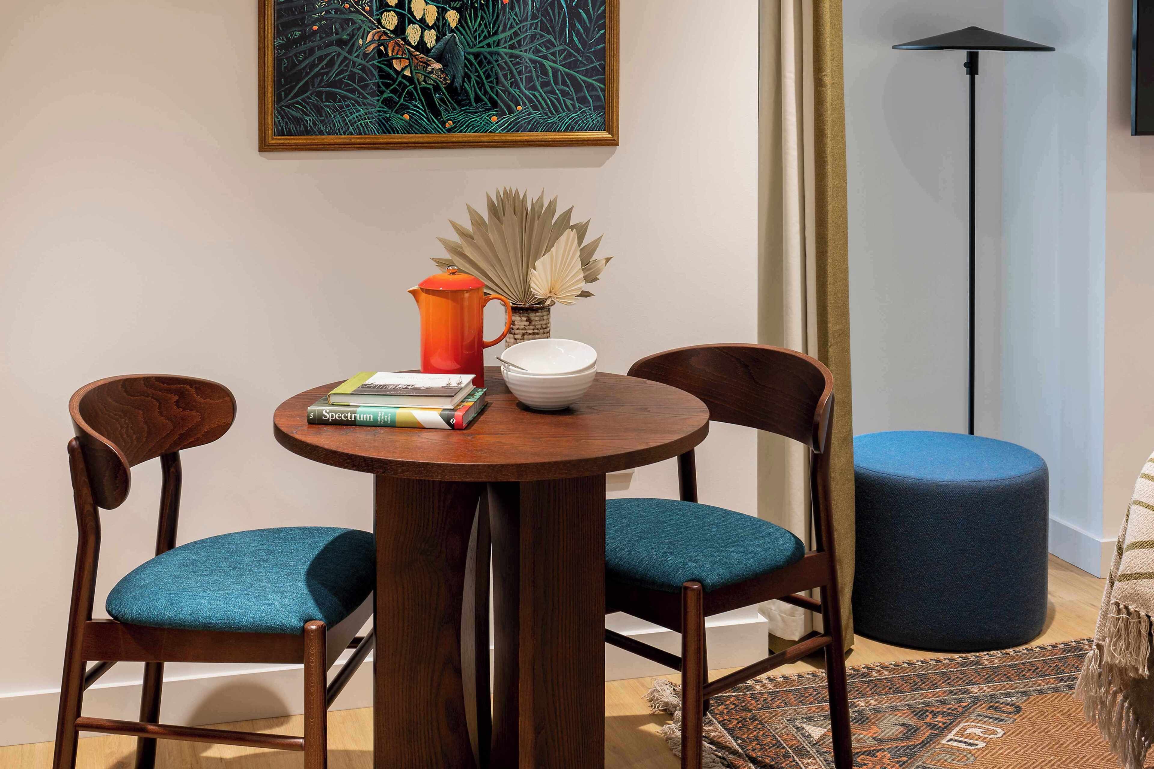 Un coin repas avec une table ronde en bois, deux chaises dotées de coussins turquoise, un petit bol blanc, un pichet rouge et un vase contenant des plantes séchées.
