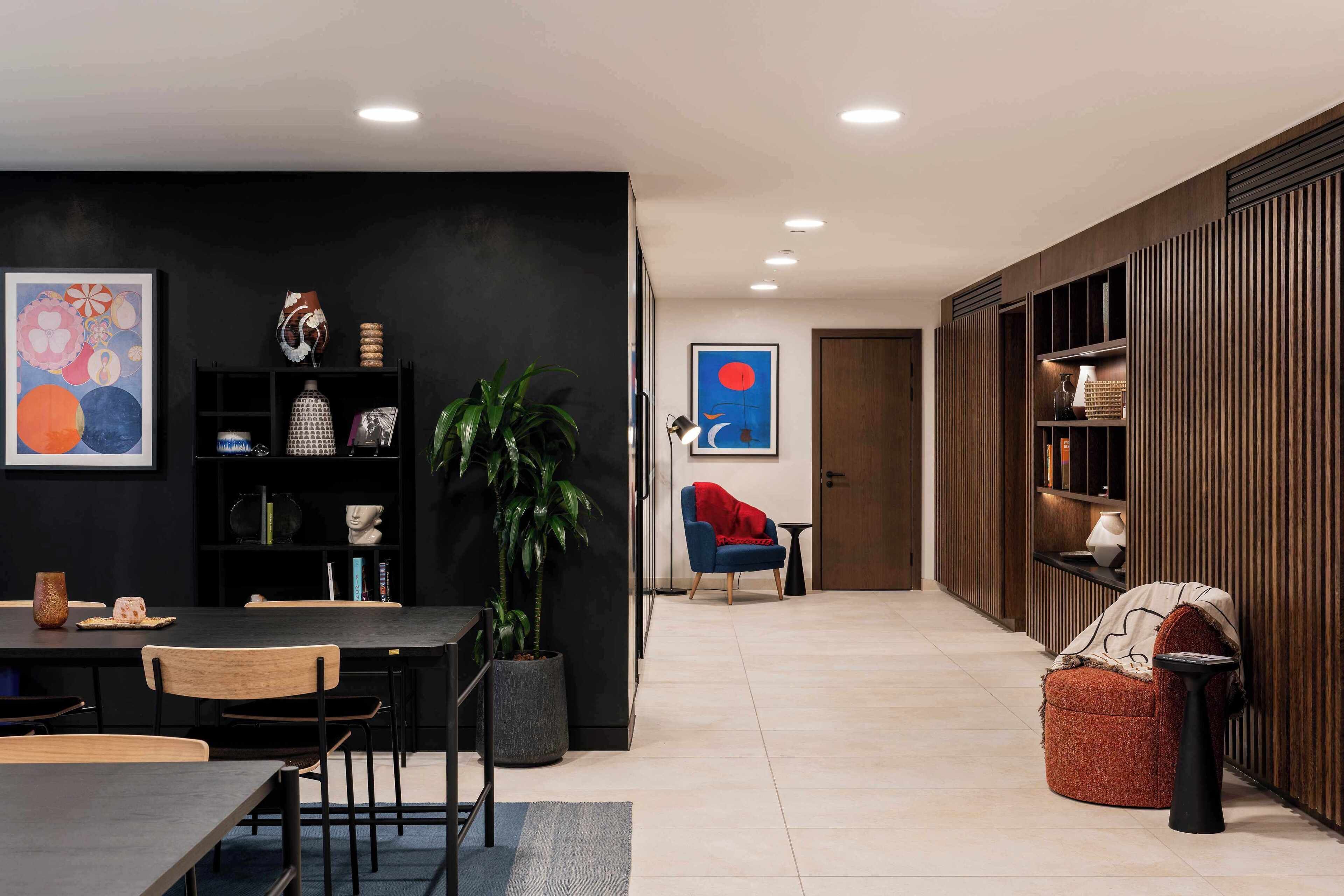 A modern living room with a dining area, featuring a black wall with artwork, a potted plant, a red armchair, and a side table.