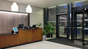 A reception area with a wooden reception desk, two pendant lights, a digital kiosk, and potted plants. - Cove Landmark Pinnacle (London)