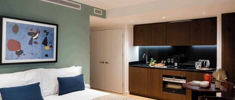 Una habitación de hotel moderna con una cama, una mesa de comedor con sillas, una zona de cocina y un cuadro abstracto enmarcado en la pared.