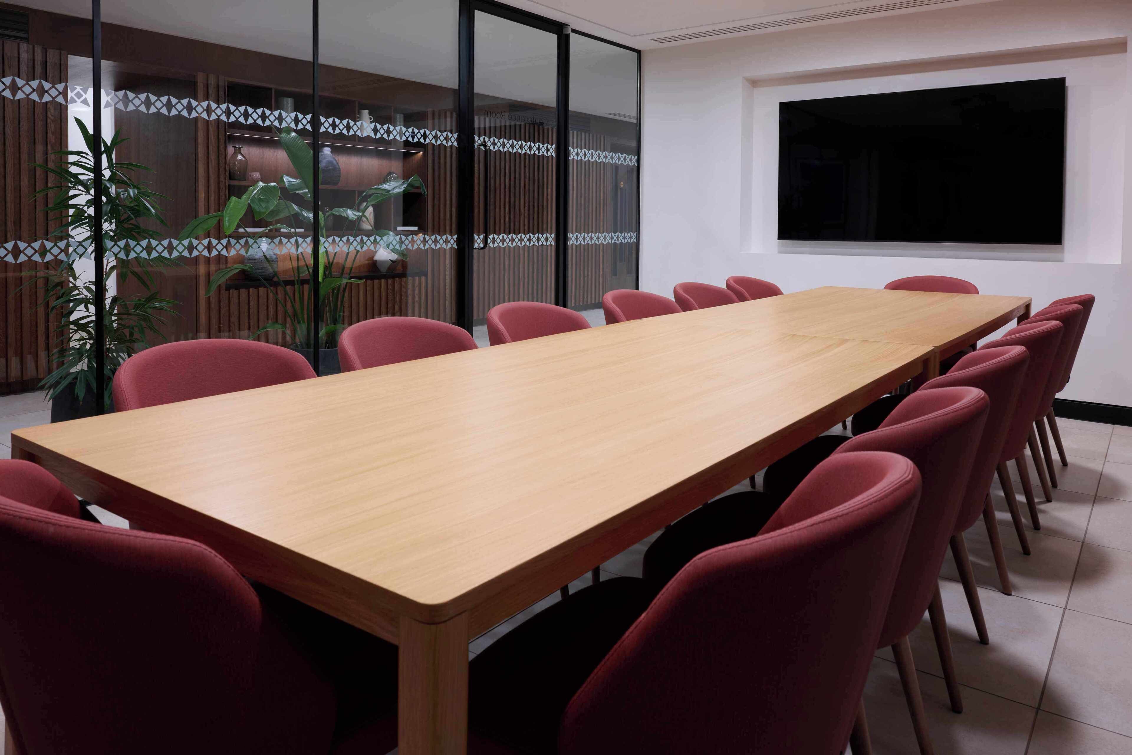 Una sala de conferencias con una larga mesa de madera, sillas rojas, un ventanal grande y un panel decorativo en la pared.