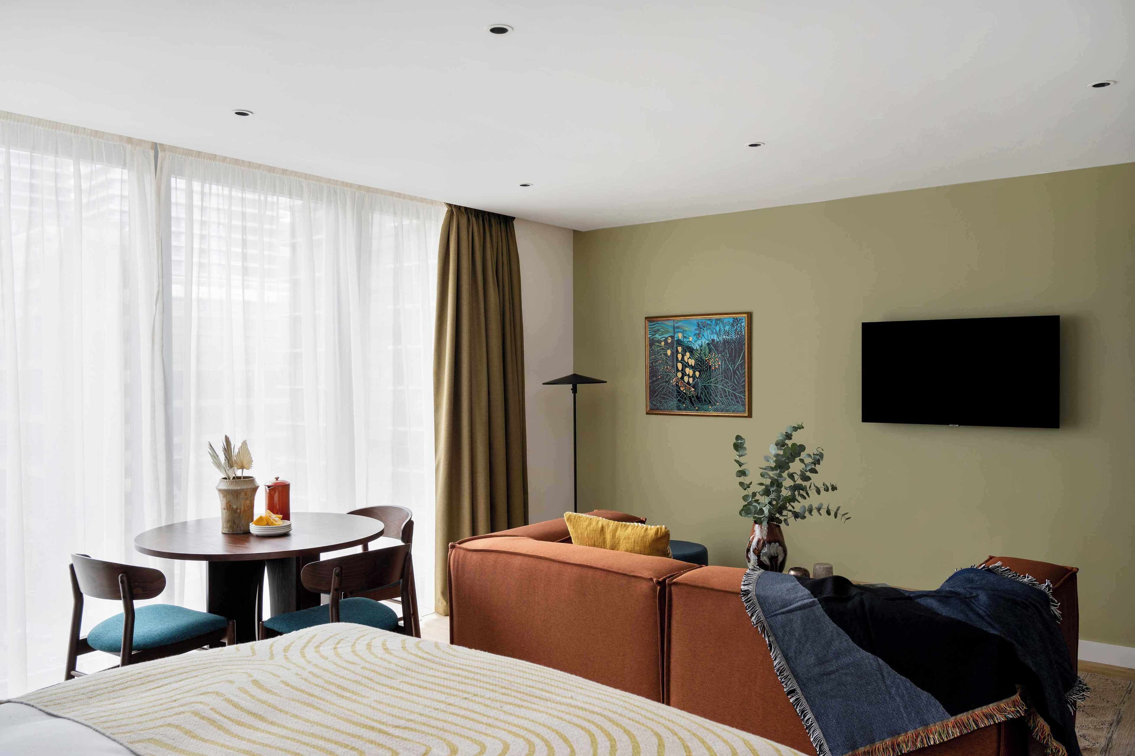 A hotel room with a bed, a sofa, a dining table with chairs, a TV, and a window with curtains.
